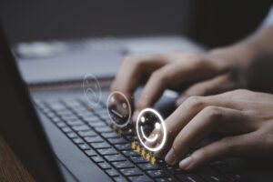 Na imagem vemos uma pessoa digitando em um notebook e aparece na acima dos dedos um emoji feliz, um emoji sem reação e um emoji triste como estrelas abaixo dos emojis.