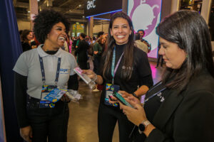 Registro do evento Conarec 2022. Três mulheres sorridentes interagindo entre si. Elas estão vestidas com roupas pretas e uma delas está com camisa branca. Uma mulher é negra e as outras duas são brancas. Elas não estão posando para a foto, é uma captura espontânea.