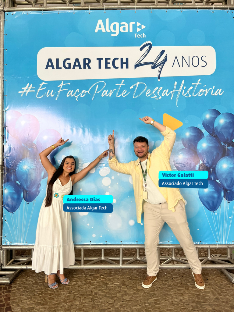 Na imagem, tem um backdrop de cor azul com o selo: Algar Tech 24 anos - escrito em fonte cor azul escuro dentro de uma barra de cor branca. Abaixo, está escrito em fonte branca e cursiva: #Eu Faço Parte Dessa História. Nas duas laterais têm aglomerados de balões flutuantes de cor azul escuro. Na frente do backdrop estão duas pessoas, associados da Algar Tech. A primeira é a Andressa Dias, uma mulher de pele branca, levemente bronzeada. Ela tem os cabelos pretos longos lisos e está sorrindo. Ela veste um vestido branco longo, com duas flores de cor azul com o miolo vermelho e verde com o miolo vermelho, bordadas na cintura. Ela está com os braços apontados para cima na direção dos textos. Ao lado dela, está o Victor Galatti. Ele é um homem de pele branca, cabelos curtos de cor castanha, tem barba e veste uma camisa branca sem estampa, uma blusa social de cor amarelo claro por cima e uma calça de cor creme. Ele está sorrindo e também aponta os braços e dedos pra cima.