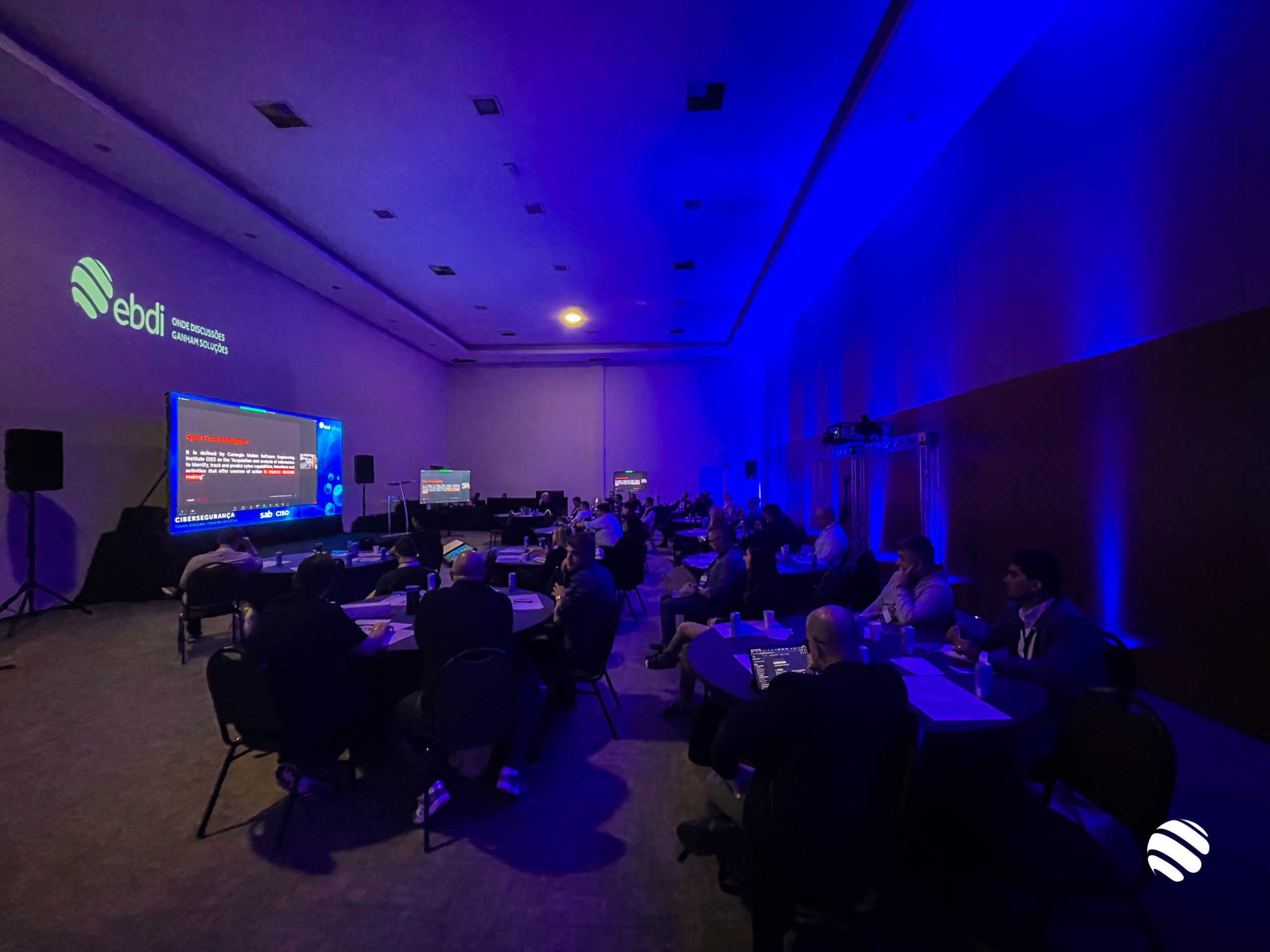 Na imagem vemos uma sala de conferencias com a iluminação baixa e levemente azulada. Mesas com algumas pessoas sentadas. 